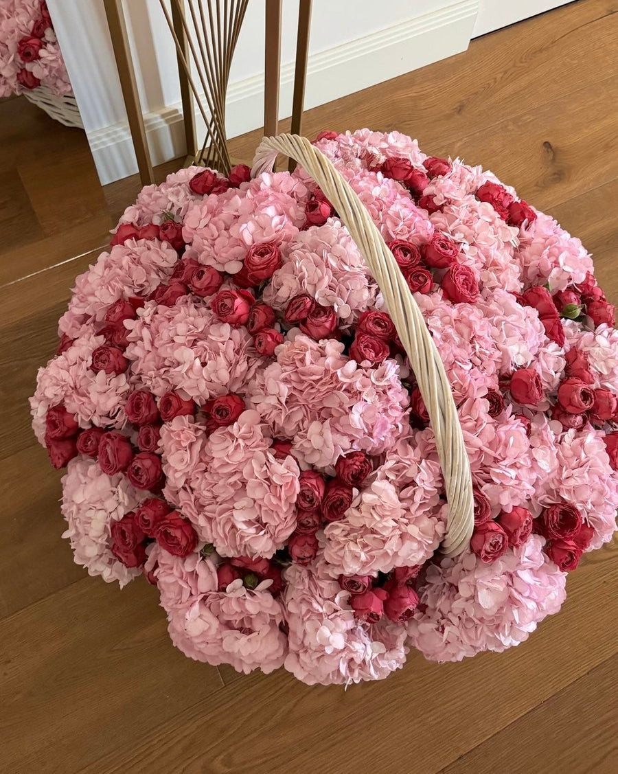 A large decorative floral arrangement in a round woven basket, featuring light pink hydrangeas and clusters of deep red roses, placed on a wooden floor.