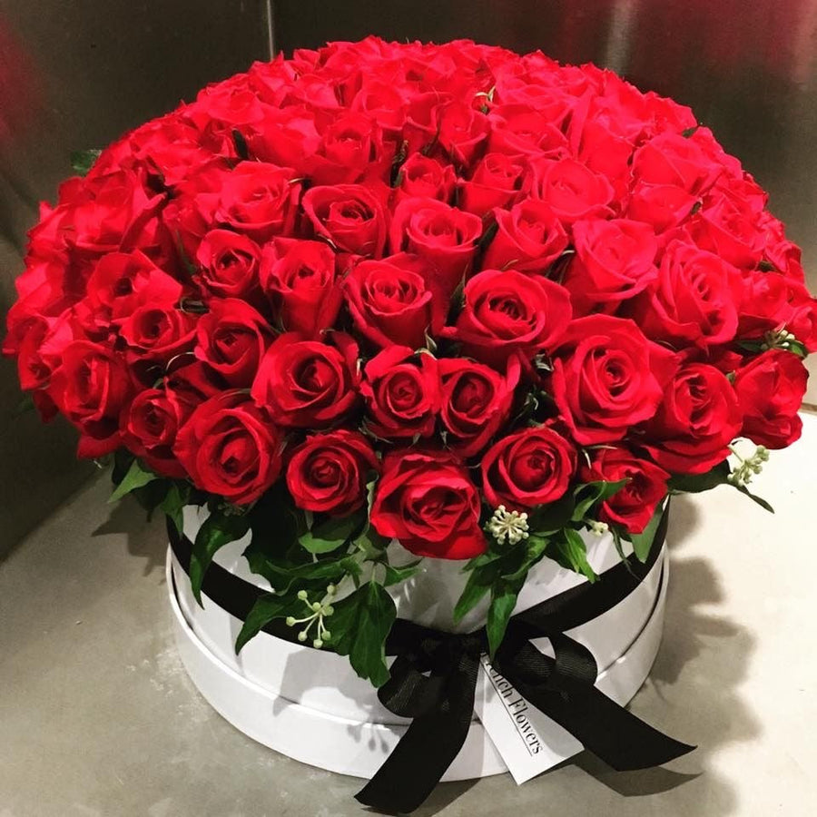 A large round bouquet of vibrant red roses arranged in a white oval box with a black ribbon and bow. A small white tag is attached to the ribbon, and the flowers are photographed against a neutral indoor background.