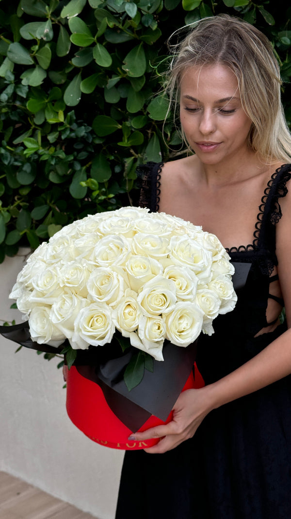 A woman in a black lace-trim dress holding a large round bouquet of white roses in a red box, posed against a green leafy wall.