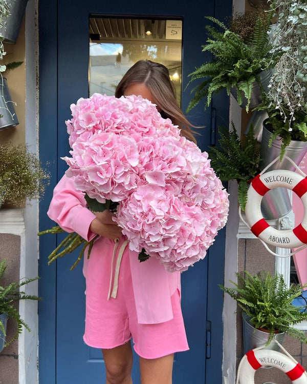A woman in a pink lounge set stands in front of a blue door holding a massive bouquet of light pink hydrangeas. The setting is a decorated storefront with potted ferns, a white life preserver sign, and warm ambient lighting.