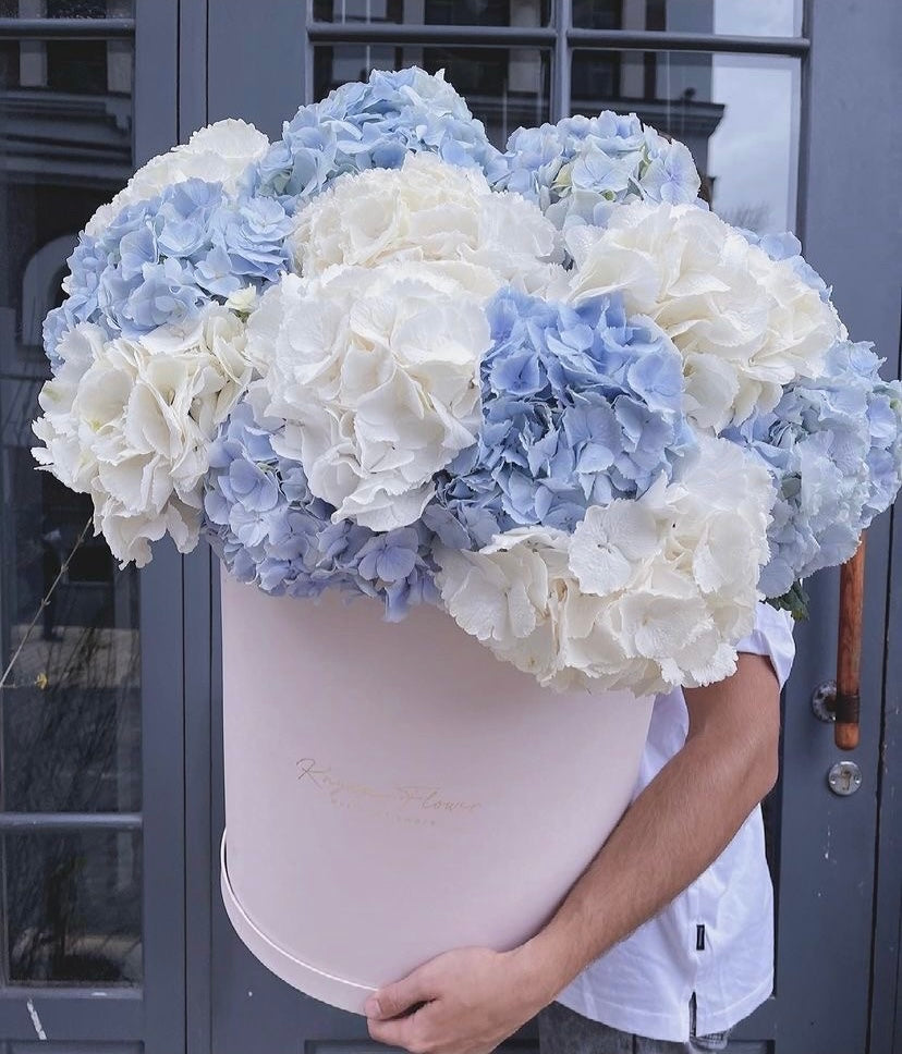 A large round pink flower box filled with pale blue and white hydrangeas, held by a person in front of a gray glass-paneled door.