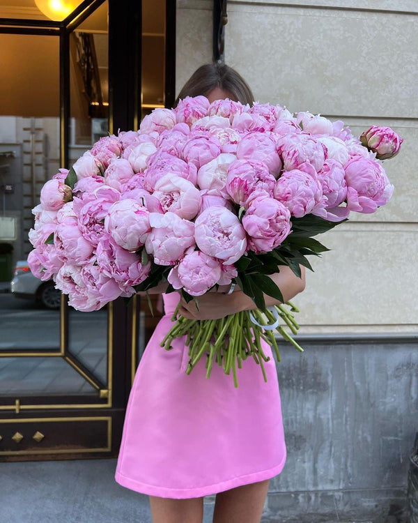 A person in a bright pink dress holds a massive bouquet of light pink peonies in front of their face, standing outdoors against a beige stone wall and a dark-framed mirror.