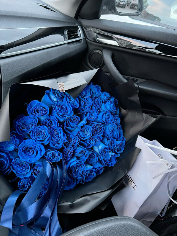 A large bouquet of vivid royal blue roses wrapped in black paper and blue ribbon, placed inside a car interior next to a white shopping bag.