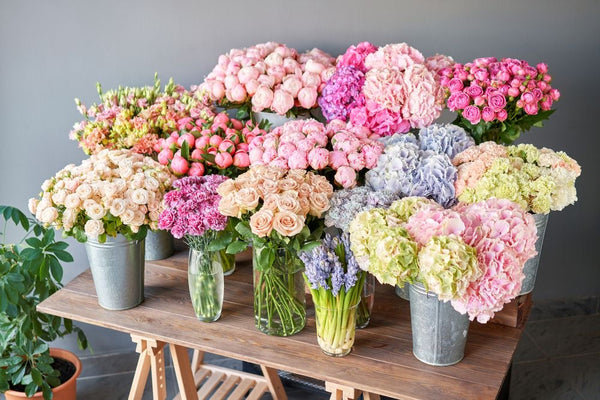 A large, vibrant floral arrangement featuring pink, peach, lavender, and light green hydrangeas, roses, and other cut flowers in galvanized metal buckets and glass vases, displayed on a wooden table against a gray wall.