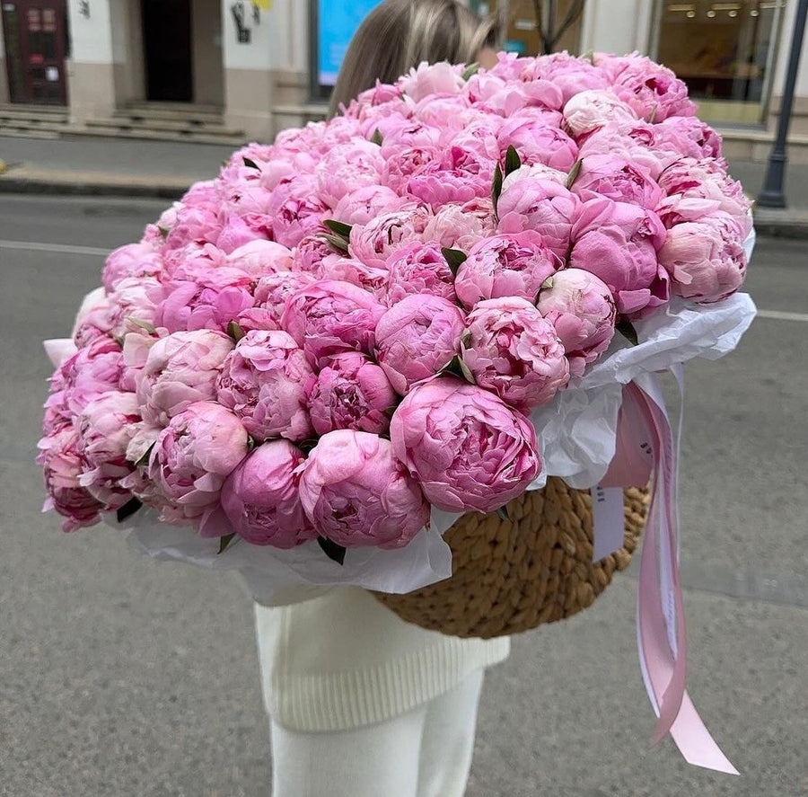 A large bouquet of pink peony flowers arranged in a round woven basket with white tissue paper and a light pink ribbon, held by a person standing outdoors on a city street.