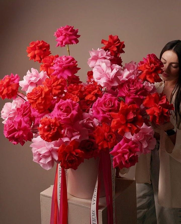A large bouquet arrangement of artificial-looking roses in shades of red, pink, and coral, displayed in a light pink vase with pink ribbons, with a woman partially visible on the right side.