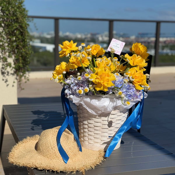 A white woven basket filled with bright yellow daffodils, small white and lavender flowers, and blue satin ribbons, placed on a dark outdoor table. A straw hat rests in front of the basket, with a balcony railing and distant cityscape in the background.