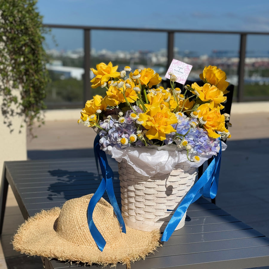 A white woven basket filled with bright yellow daffodils, small white and lavender flowers, and blue satin ribbons, placed on a dark outdoor table. A straw hat rests in front of the basket, with a balcony railing and distant cityscape in the background.