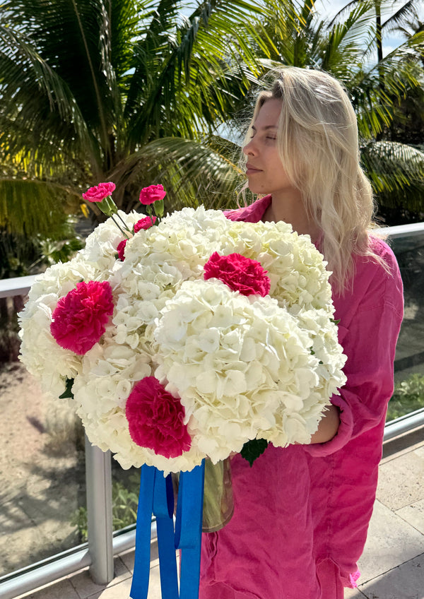 A woman in a bright pink jacket holds a large bouquet of white hydrangeas with bright pink carnations and blue ribbon accents, set against a tropical background of palm trees.