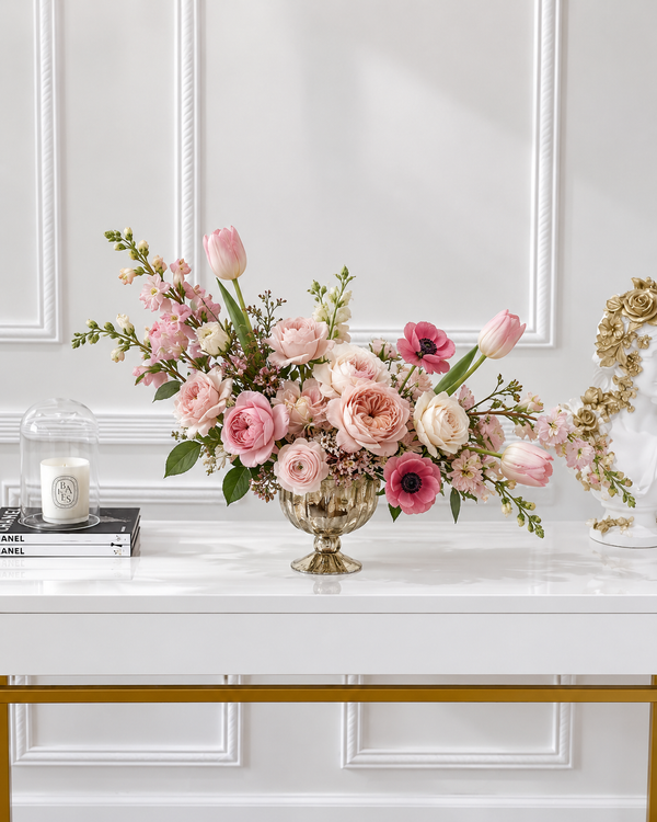A pink and white floral arrangement in a gold pedestal vase on a white console table, by Mia Mur Miami.
